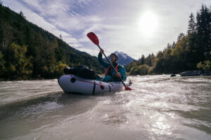A serene view of a calm river surrounded by lush greenery, with a person in a packraft paddling under a clear blue sky. Ideal for showcasing beginner-friendly packraft trails in Europe.